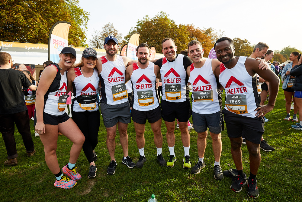 A group of seven runners wearing Shelter vests stand in a semi-circle with their arms around each other before the race starts