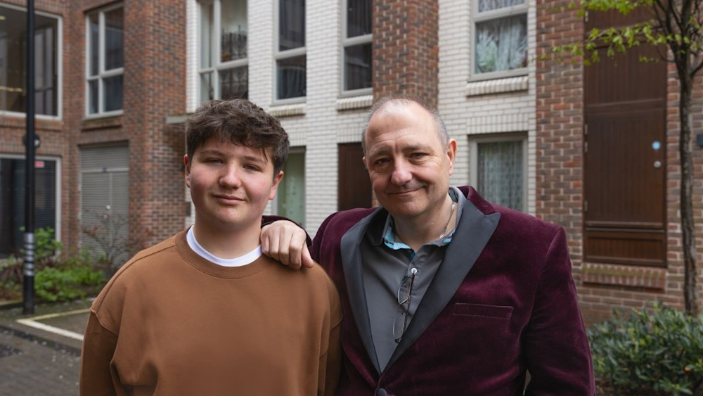 A middle-aged man wearing a red velvet jacket, and his teenage son stand in front of a block of well-maintained social housing.