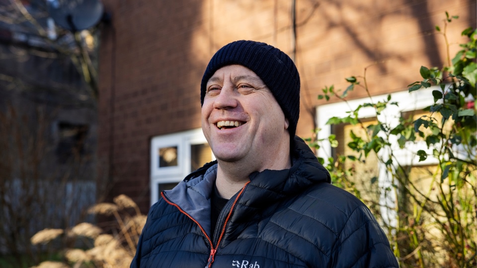A man dressed in winter clothes, smiling outside a council house.