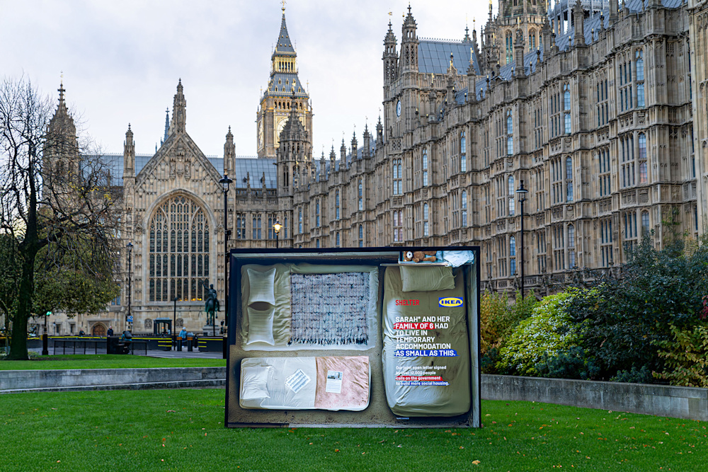 A large Shelter and IKEA poster outside the Houses of Parliament. It shows a plan of a tiny room with three small beds. Text reads 'Sarah and her family of six had to live in temporary accommodation as small as this'.