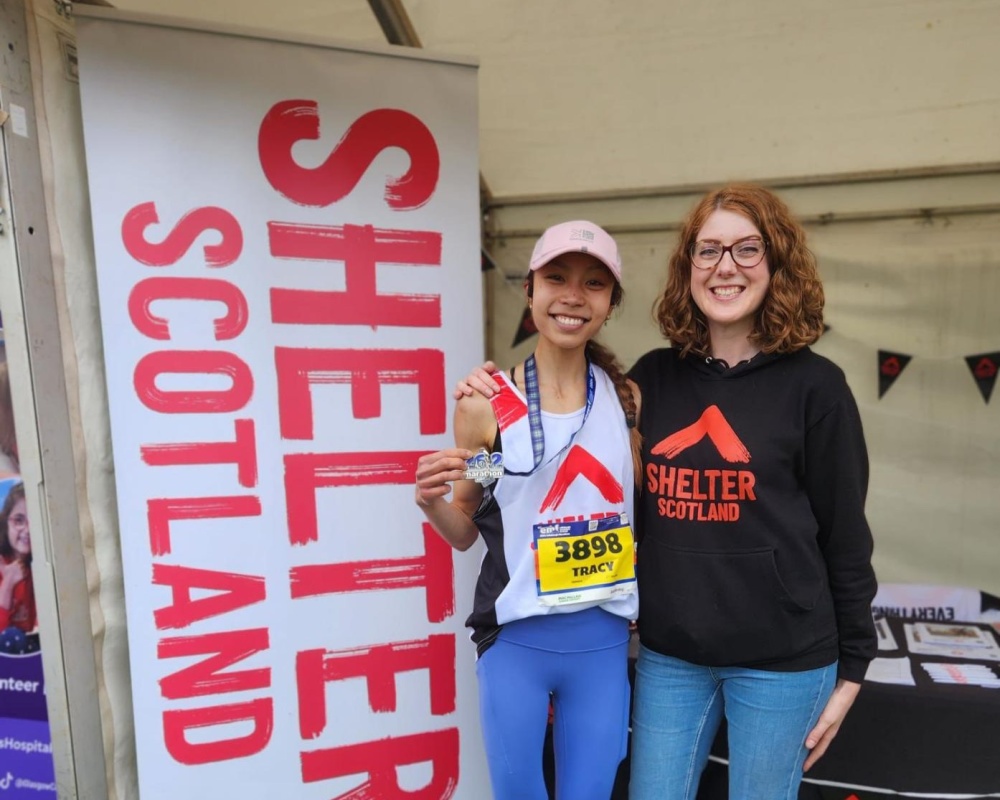 A smiling marathon finisher holds up her medal next to a Shelter Scotland banner. Beside her is a member of the Events team wearing a Shelter Scotland hoodie.