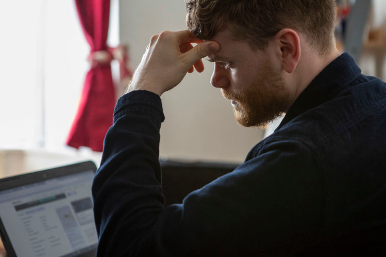 A man sitting at a computer looking through rental listings. He appears stressed with his head in his hand.