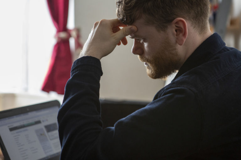 A man sitting at a computer looking through rental listings. He appears stressed with his head in his hand.