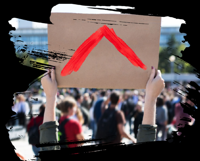 Close up of a person's hands and arms, holding a cardboard sign painted with the red Shelter logo. A crowd of demonstrators is in the background.