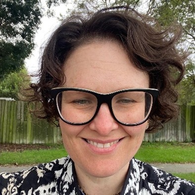 The face of a woman who is of White ethnicity with short, brown hair wearing square-framed, black glasses. She is smiling and a park is in the background.