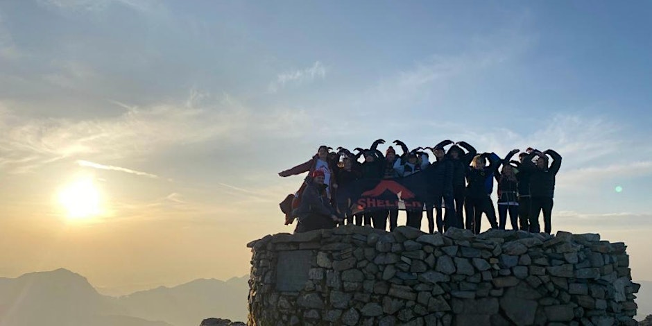 A group of walkers standing on a rocky crag holding a Shelter banner, while the sun rises behind them. They are holding their arms up in a triangle shape to mimic the Shelter logo.