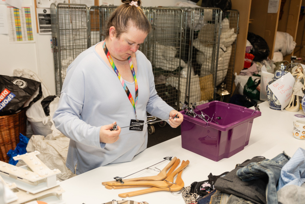 Behind the scenes at a Shelter shop, a volunteer selects clothes hangers from a plastic box. She is a white woman wearing a grey jumper and rainbow lanyard.