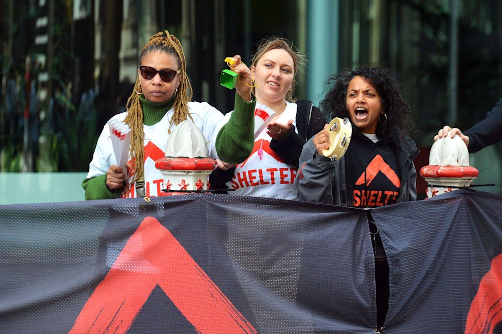 Three people wearing Shelter hoodies at the cheer point of a race, standing behind a Shelter banner. They are clapping, shaking tambourines and calling encouragement to passing runners.