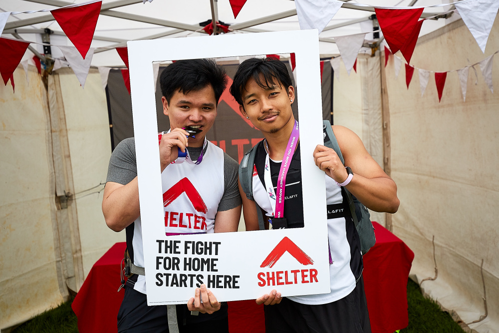 Two men wearing Shelter vests and medals hold up a frame that reads 'The fight for home starts here'.