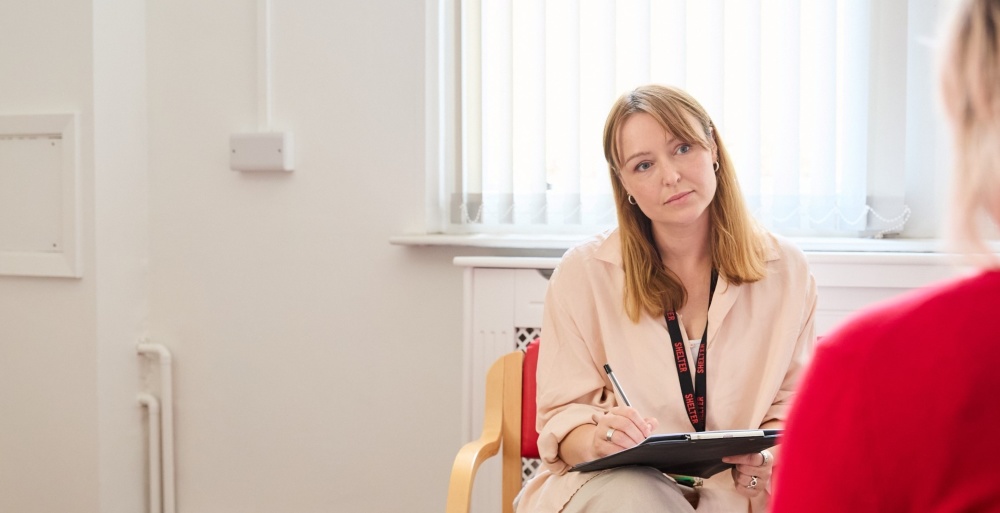 A woman with her back to the camera speaks to a Shelter adviser, who listens attentively while writing on a clipboard.