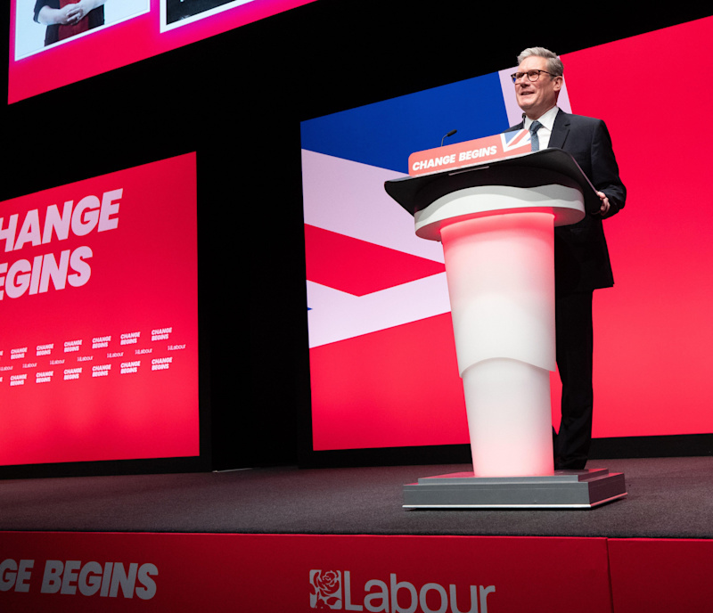 Prime Minister Keir Starmer on stage in front of a white lectern, in front of a red projection which reads 'change begins'. 