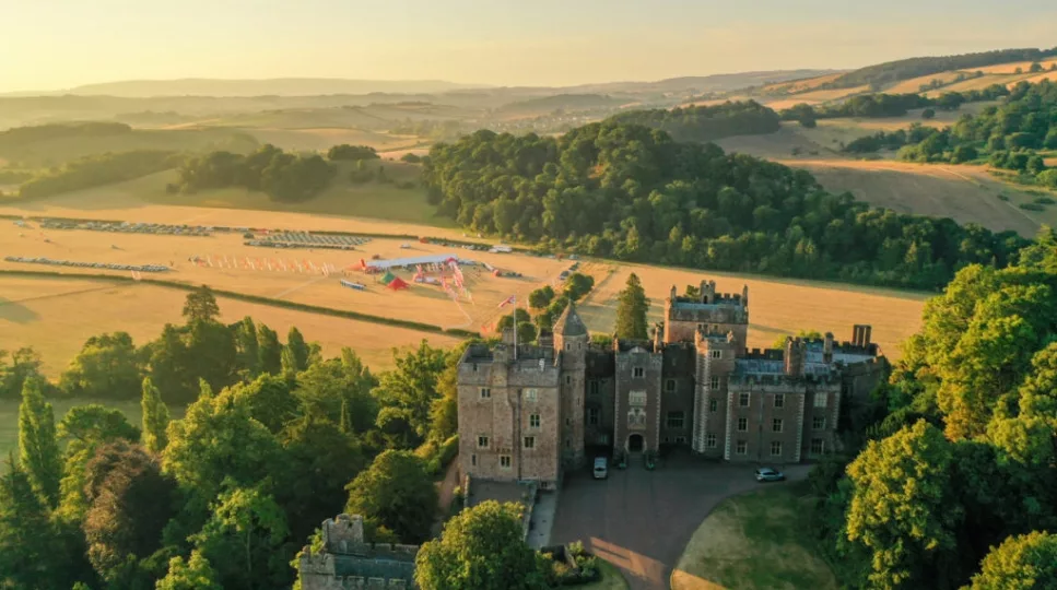 A hilltop view of an impressive castle surrounded by trees and fields. Next to the castle a camp is set up with colourful tents.
