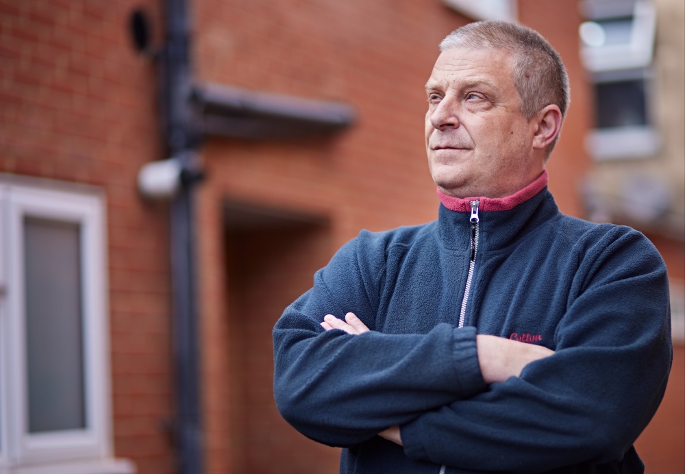 A man wearing a blue fleece stands outside a block of flats with his arms folded.