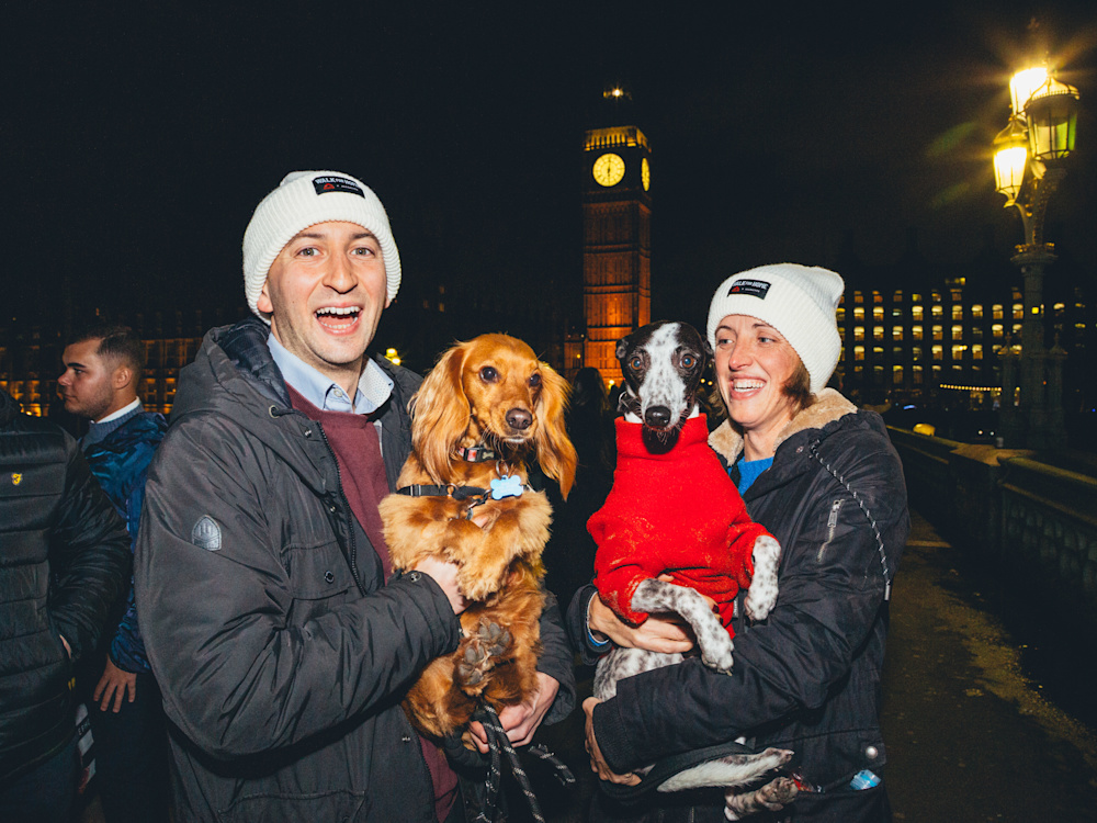 Two people hold their dogs at a walk for home event in London, wearing Shelter hats.
