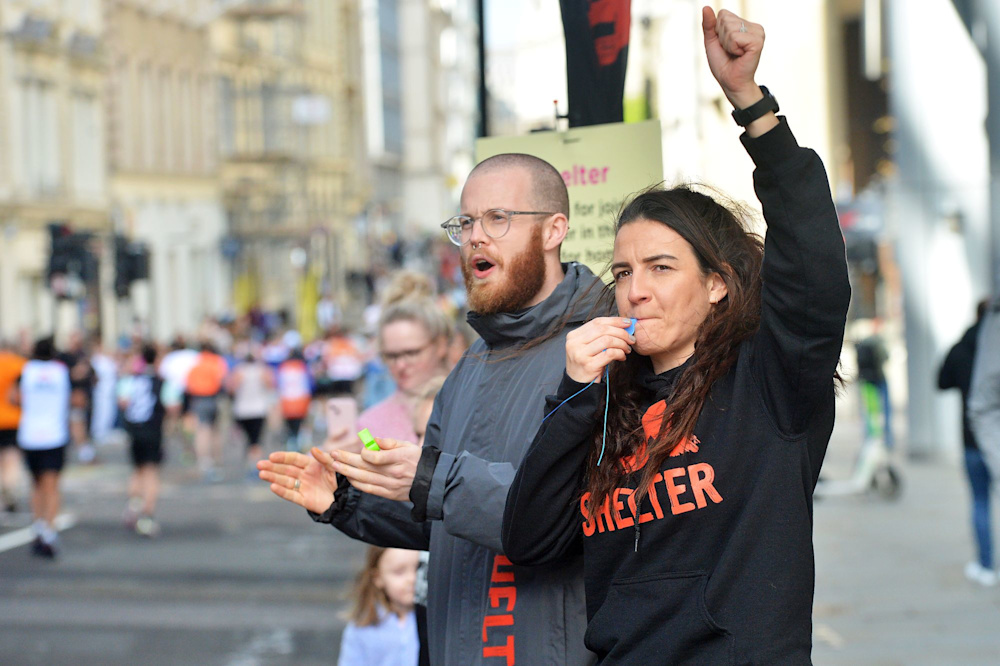 Two members of the Shelter Events team at the cheer point of a race, wearing Shelter coats and hoodies. One person is clapping and the other is holding a whistle.