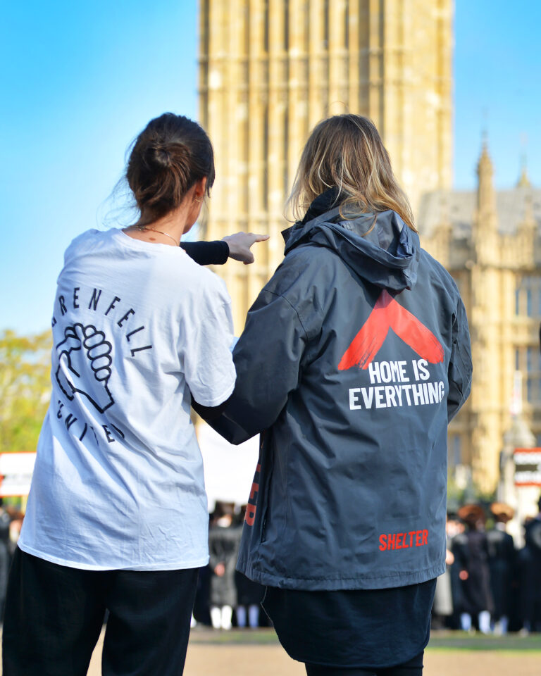 Two campaigners talk next to Big Ben. One wears a Grenfell United T shirt and one wears a Shelter raincoat that reads 'Home is everything'.