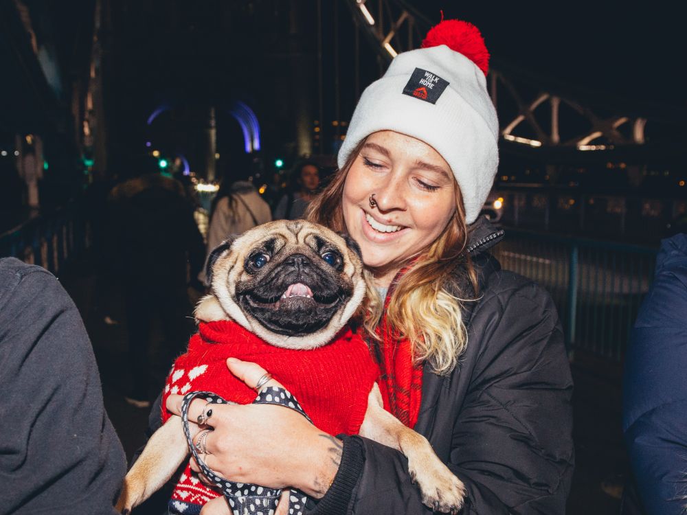 A woman wearing a red and white bobble hat holds up an adorable pug in a red dog jumper.