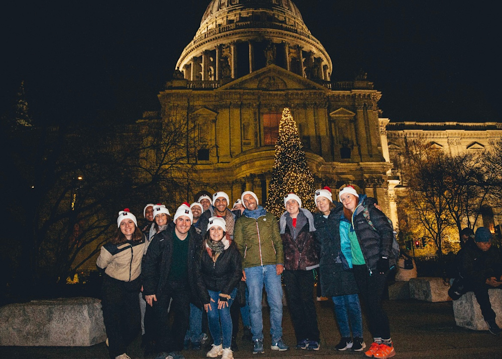 A group of people wearing red and white Walk for Home bobble hats pose for a photo by a huge Christmas tree in London.