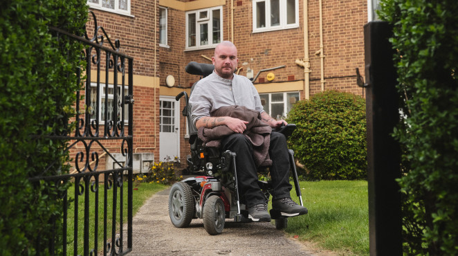 Man looking defiant sits in a wheelchair in front of a block of flats