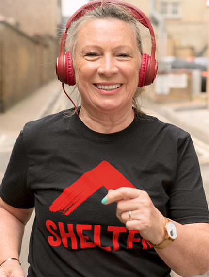 A woman wearing headphones and a T-shirt with a Shelter logo is smiling as she runs down a city street