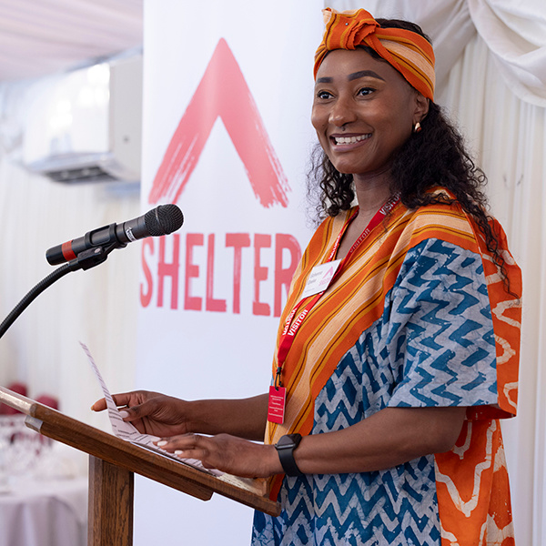 Peer researcher Uche smiles as she stands at a podium by a Shelter logo.