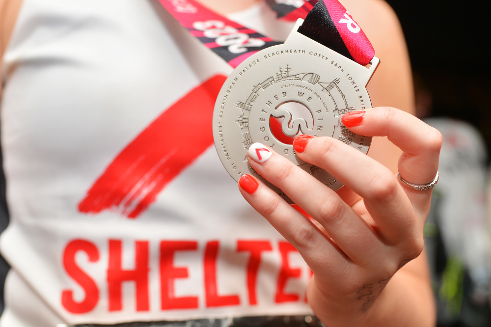 Close-up of a woman's hand holding the London Marathon finisher's medal. Her nails are painted red, with the Shelter logo on the index finger.