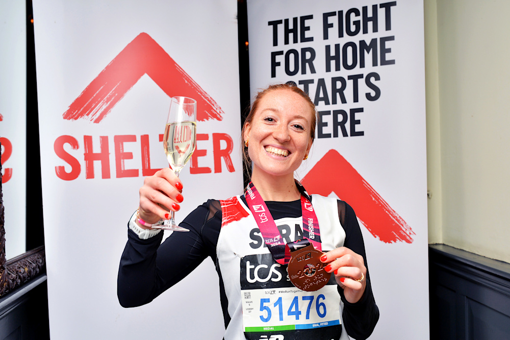 A woman in a Shelter vest celebrates after finishing the marathon. She holds up her medal and a glass of champagne. In the background is a banner than reads 'The fight for home starts here'.