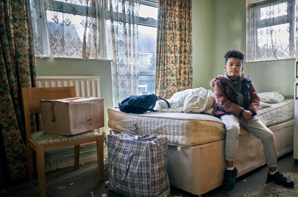 A young boy sits on a mattress in a dirty room filled with bags and a box