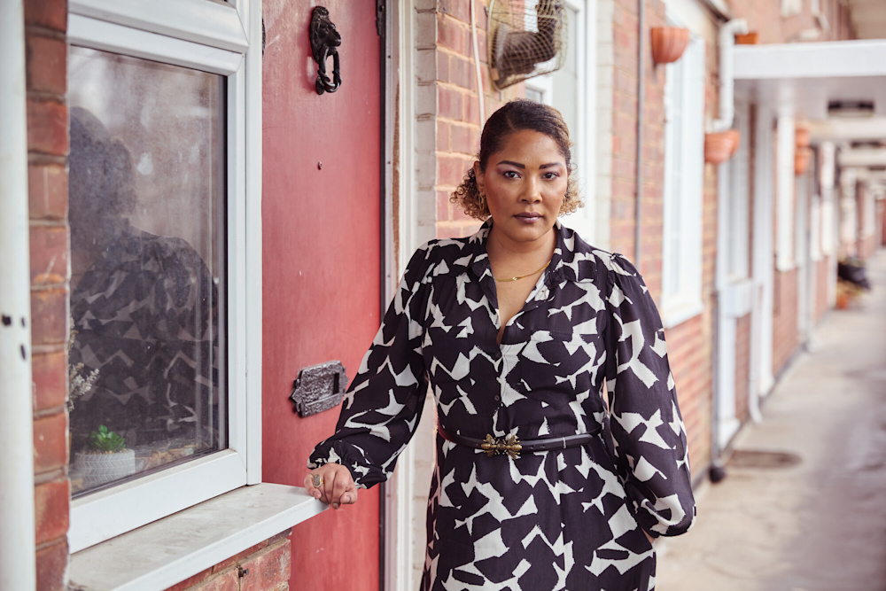 A woman Sireena in a black and white dress leans on the red door of her social home within a block of flats.