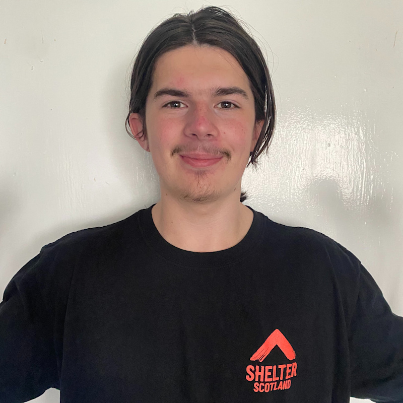 A man of White ethnicity, with brown hair, a moustache and goatee stands in front of a white wall. He is smiling and wearing a black t-shirt with the Shelter Scotland logo on the left-hand side of his chest.