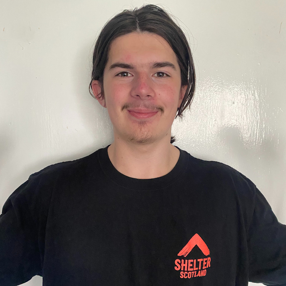A man of White ethnicity, with brown hair, a moustache and goatee stands in front of a white wall. He is smiling and wearing a black t-shirt with the Shelter Scotland logo on the left-hand side of his chest.