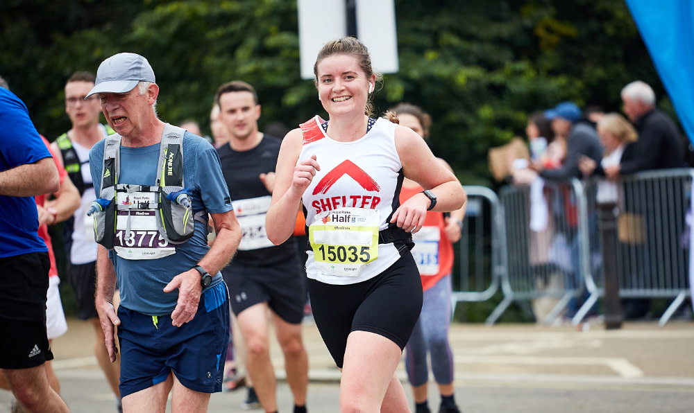 A woman in a white Shelter jersey running and smiling at the camera during the Royal Parks Half Marathon race