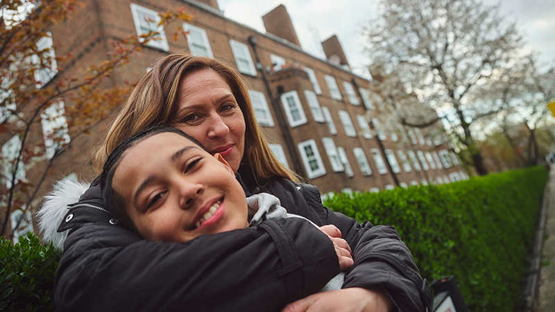 A mother and teenage son are smiling as they embrace outside a block of flats. 