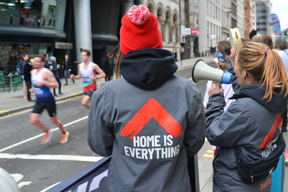 Two women wearing Shelter jackets stand with their backs to the camera, cheering on runners from the sideline. One is wearing a bright red bobble hat and the other shouts into a megaphone.