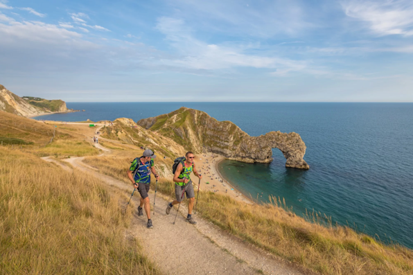 Two people walking along a cliffside path, with a beautiful view of the sea and rock formations behind them.