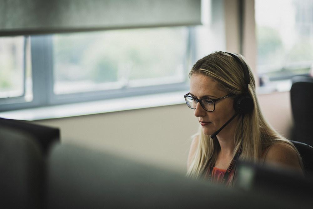 A helpline advisor wearing a headset and glasses sits down facing a computer. 