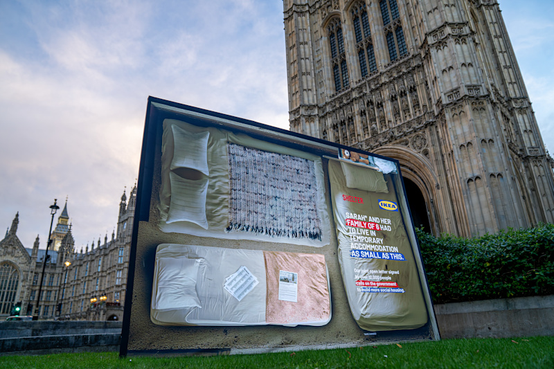 A large Shelter and IKEA poster outside the Houses of Parliament. It shows a plan of a tiny room with three small beds. Text reads 'Sarah and her family of six had to live in temporary accommodation as small as this'.