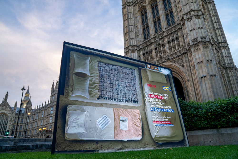 A large Shelter and IKEA poster outside the Houses of Parliament. It shows a plan of a tiny room with three small beds. Text reads 'Sarah and her family of six had to live in temporary accommodation as small as this'.