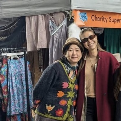 Two women are stood side by side at a market stall, smiling. On the left is a Shelter staff member with black hair. She is wearing a hat and blue cardigan with floral detail. Tassia has long brown hair. She is wearing tinted glasses and a red coat. 