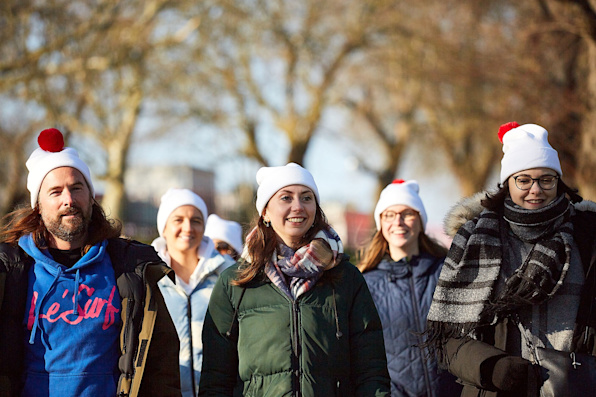 A group of five people walking in a park, wearing coats and bobble hats.