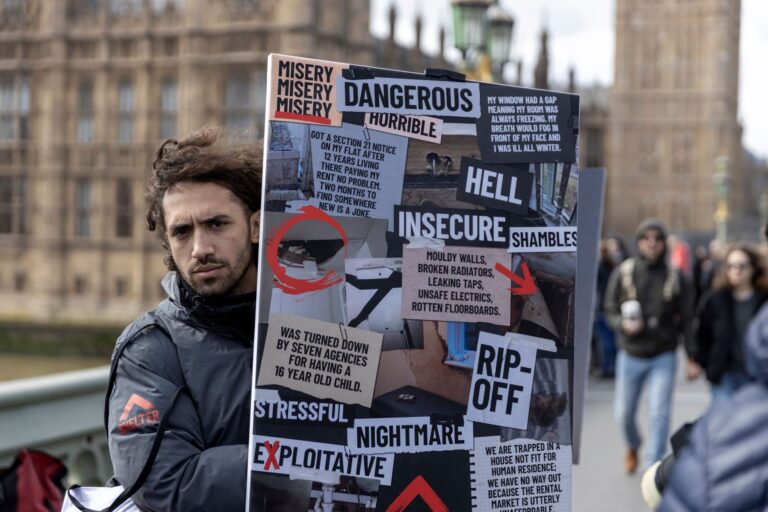 A man in a black Shelter-branded jacket stands outside the House of Parliament in London, holding a placard with a collage of words and images related to renting.