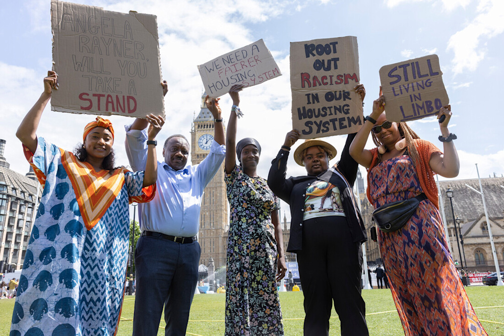 A group of people stand outside Big Ben, Westminster, London. They hold various signs demanding an end to racism in our housing system.