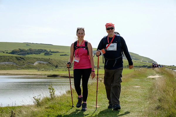 Two hikers carrying walking poles smile as they walk past a lake on a sunny day