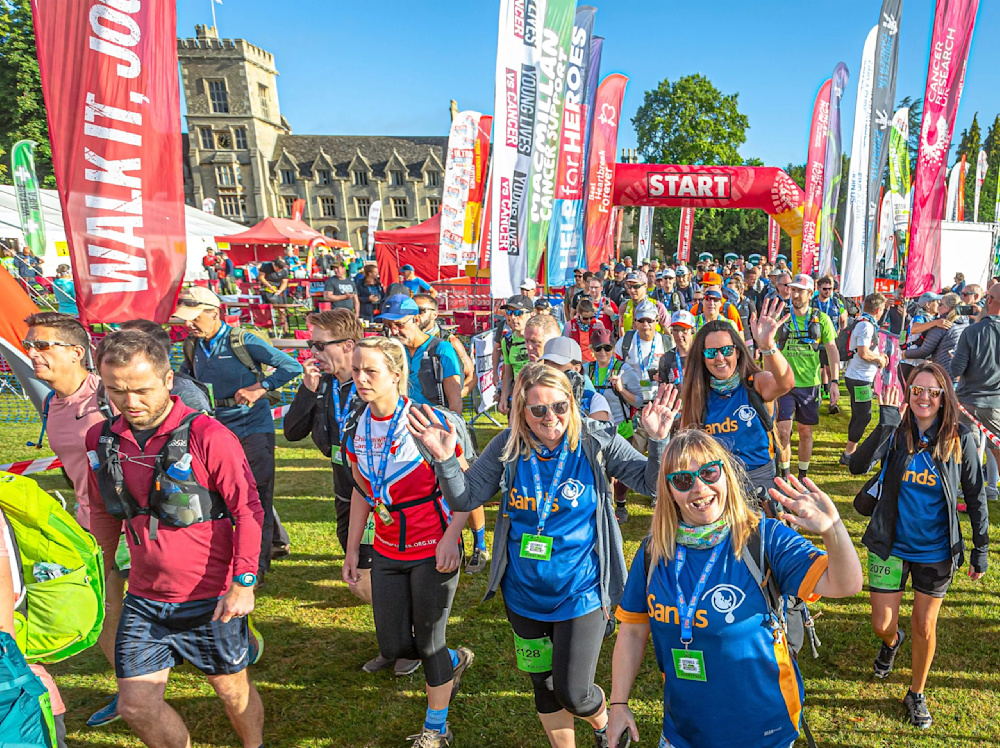 A big crowd of walkers set out from the start line of the Cotswold Way challenge, with colourful banners on either side. They are waving cheerfully at spectators.