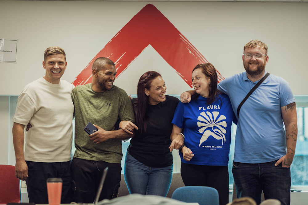 A group of Shelter staff stand in a line with their arms around each other, talking and laughing. The Shelter logo is painted on a wall in the background.