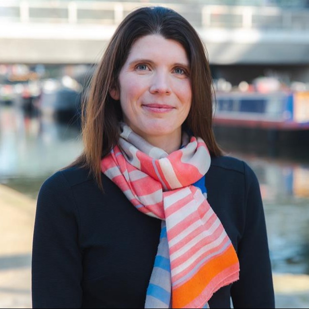 Sarah Elliott, Shelter's CEO, stands smiling in front of a canal. 