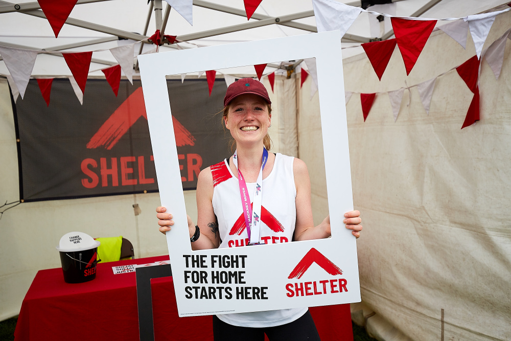 A woman wearing a medal and Shelter vest celebrates after her race. She is holding a frame that reads 'The fight for home starts here'.