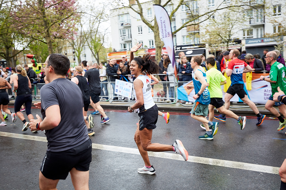 A woman runner passes the cheer point at the London Marathon and waves. She is wearing a Shelter vest that reads 'Home is everything' on the back.