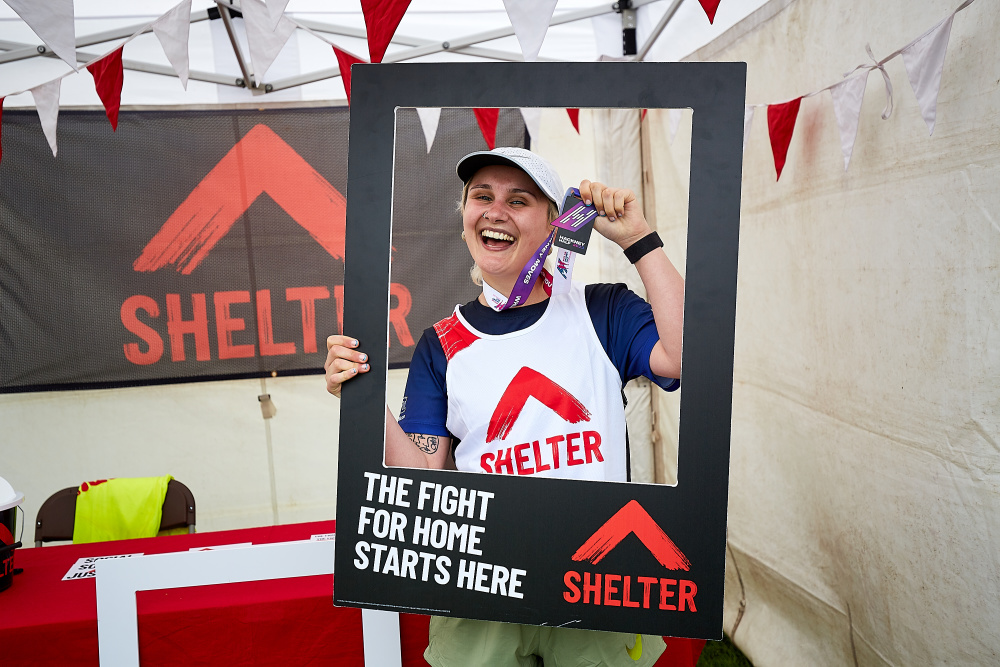 A woman in a Shelter vest triumphantly shows her medal. She is holding a frame that reads 'The fight for home starts here'.