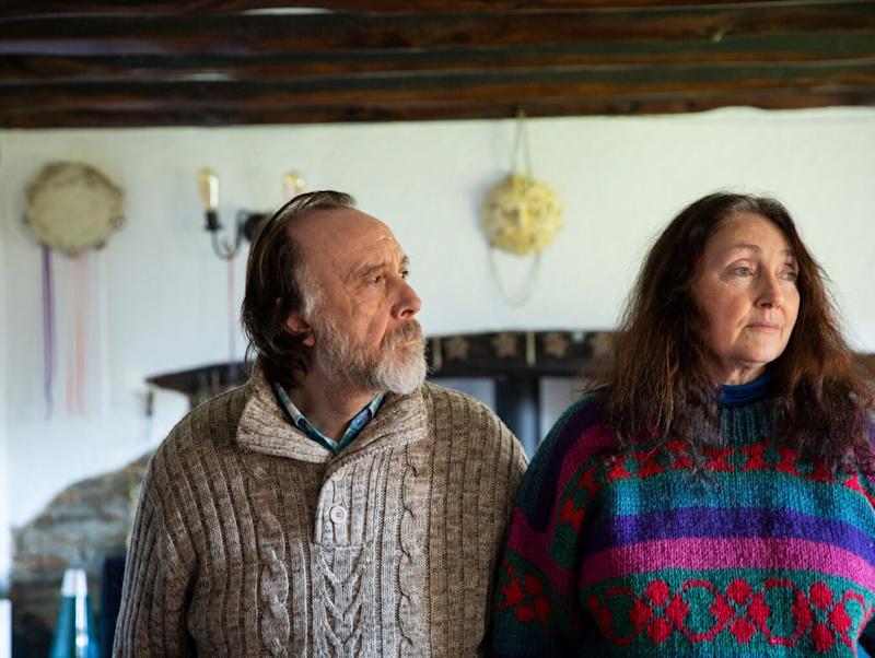 An older couple wearing jumpers stand together in their home, looking off-camera